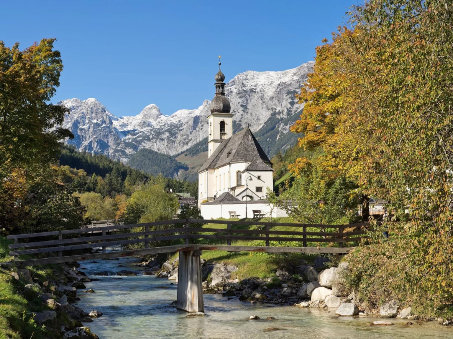 Die weiße Pfarrkirche St. Sebastian in Ramsau bei Berchtesgaden mit Zwiebelturm vor schneebedeckten Gipfeln der Reiter Alm, davor ein Gebirgsbach mit Holzbrücke und herbstlich gefärbte Bäume.