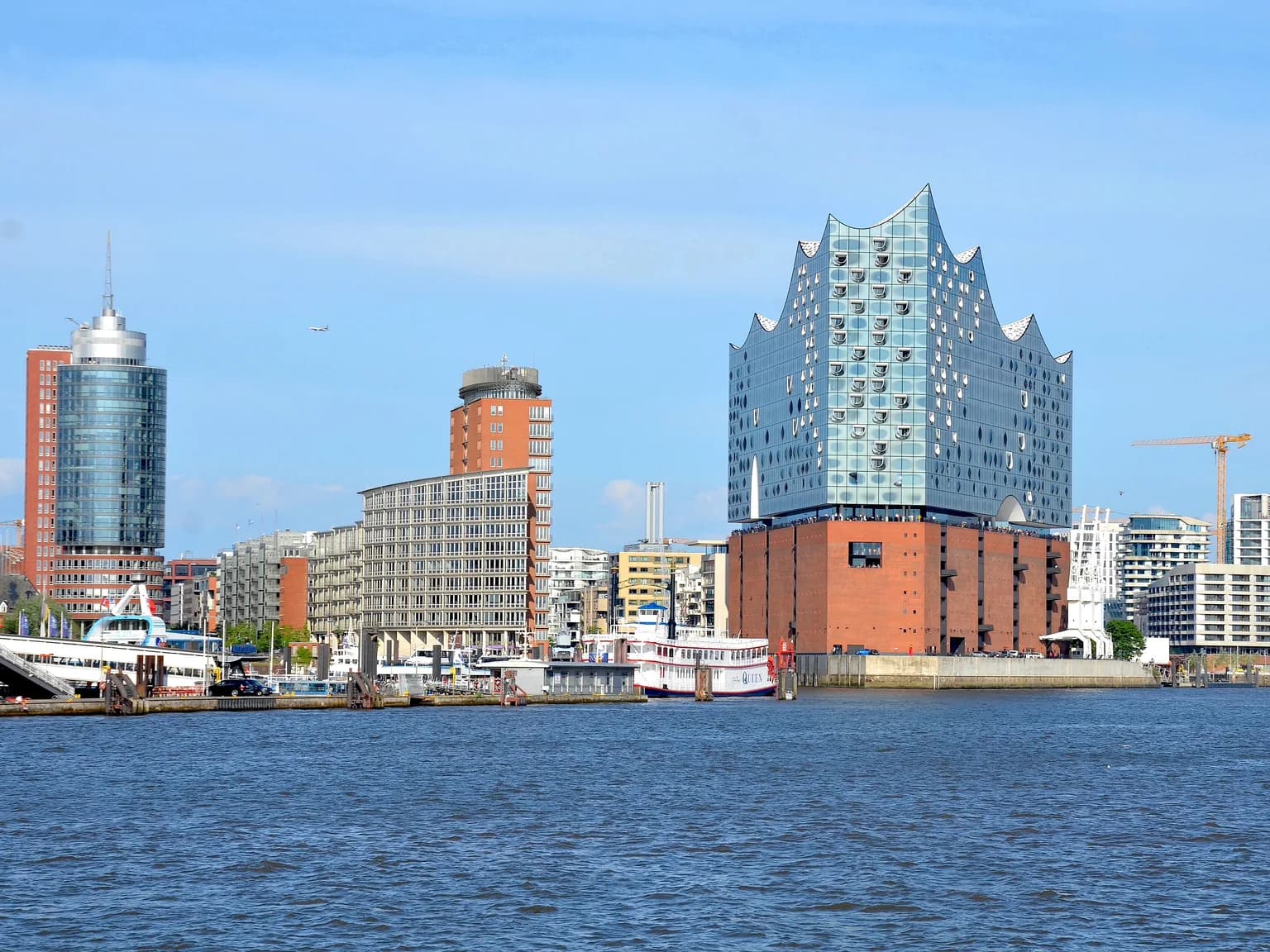 Die Elbphilharmonie mit ihrer wellenförmigen Glasfassade auf dem roten Backsteinsockel, links daneben das Hanseatic Trade Center und das Columbushaus, von der Elbe aus bei blauem Himmel gesehen.