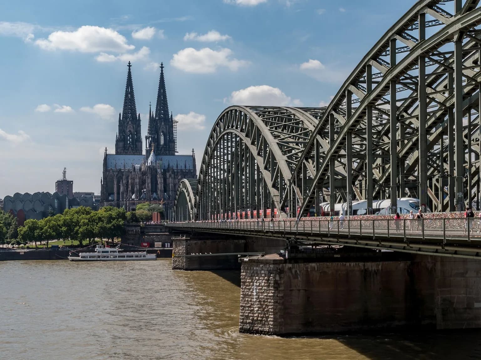 Der Kölner Dom mit seinen beiden Türmen hinter den Stahlbögen der Hohenzollernbrücke, darunter der Rhein mit einem Ausflugsschiff an einem sonnigen Sommertag.