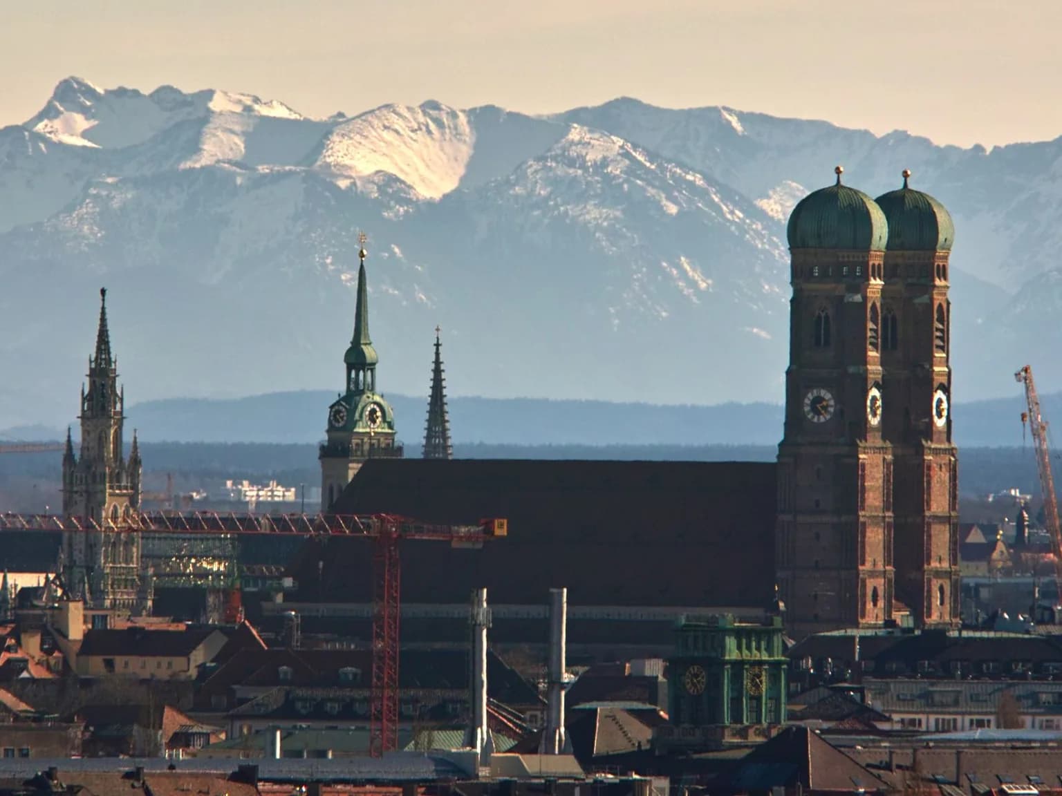 Blick über die Münchner Altstadt mit den Doppeltürmen der Frauenkirche, dem Turm von St. Peter und dem Rathausturm vor der schneebedeckten Alpenkette.