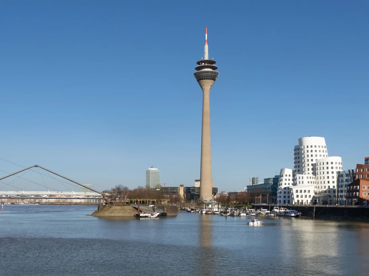 Der Düsseldorfer Rheinturm ragt bei blauem Himmel über das Hafenbecken des Medienhafens, rechts stehen die geschwungenen weißen Gehry-Bauten am Neuen Zollhof, links die Hafenbrücke.