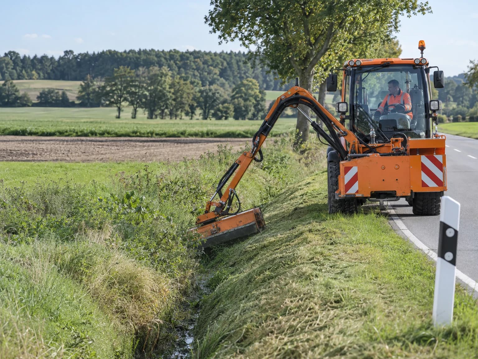 Traktor mit Auslegermäher pflegt Bankett und Graben an einer Landstraße